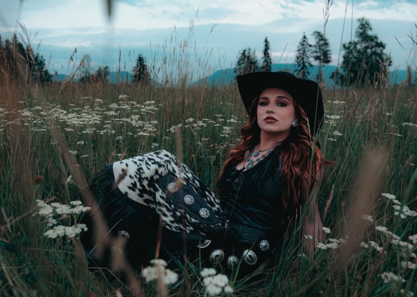 A woman with auburn hair wearing a black dress and cowboy hat sat in a fieldx